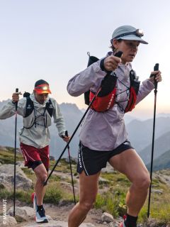 Deux randonneurs avec des lampes frontales et des bâtons de trekking gravissent un sentier de montagne rocailleux à l'aube, en vêtements de sport et avec des sacs à dos, avec les montagnes en arrière-plan.