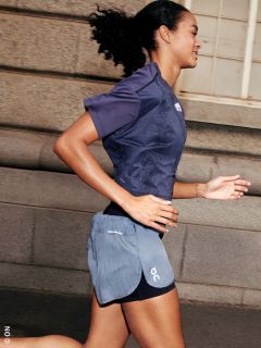 Une femme fait du jogging en plein air en tenue de sport, sourit et passe devant un bâtiment en pierre avec des fenêtres.