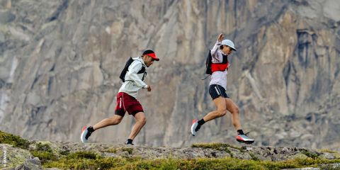 Deux personnes faisant du trail en terrain rocailleux avec une grande paroi rocheuse en arrière-plan, toutes deux avec des sacs à dos, des casquettes et des vêtements de sport.