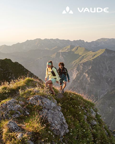 Deux randonneurs avec des sacs à dos escaladent une crête herbeuse au lever du soleil, avec des montagnes escarpées en arrière-plan. Le logo Vaude apparaît dans le coin.