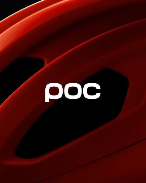 Une personne portant un manteau d'hiver orange et un casque se tient au sommet d'une montagne enneigée et regarde les montagnes enneigées au loin. Le logo POC est visible sur le casque et dans le coin supérieur droit de l'image.