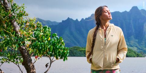 Une femme vêtue d'une veste beige se tient au bord d'un lac avec des montagnes en arrière-plan, les yeux fermés et le visage tourné vers le soleil, à côté d'un arbre feuillu.