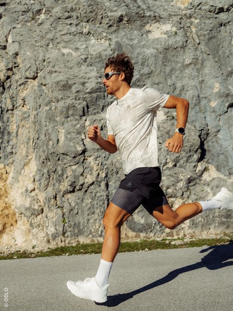 Un homme portant des lunettes de soleil et des vêtements de sport marche sous une lumière vive le long d'une route qui longe une paroi rocheuse.