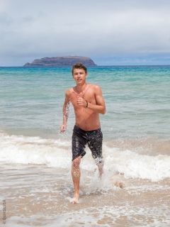 Ein hemdsärmeliger Mann mit Tattoos und schwarzen Badeshorts steht bei Sonnenuntergang im flachen Meerwasser und schaut in die Kamera. Hinter ihm rollen die Wellen heran.