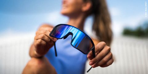 Une femme vêtue d'un haut bleu tient des lunettes de soleil de sport à effet miroir face à l'appareil photo, l'arrière-plan extérieur est flou.