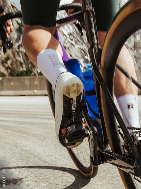 Gros plan du pied d'un cycliste sur une pédale, avec une chaussure de vélo blanche, une gourde bleue et un mouvement flou sur une route.