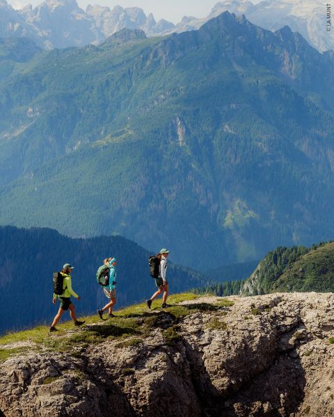 Trois randonneurs avec des sacs à dos marchent le long d'une crête rocheuse avec de hautes montagnes boisées en arrière-plan sous un ciel clair.