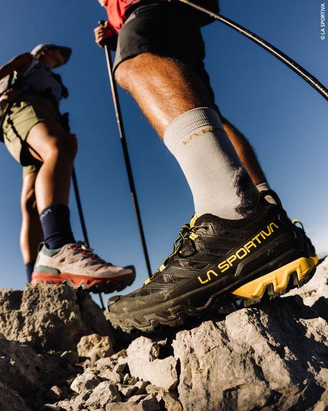 Gros plan de deux randonneurs debout sur un terrain rocheux, regardant leurs chaussures de trail La Sportiva sous un ciel bleu clair.