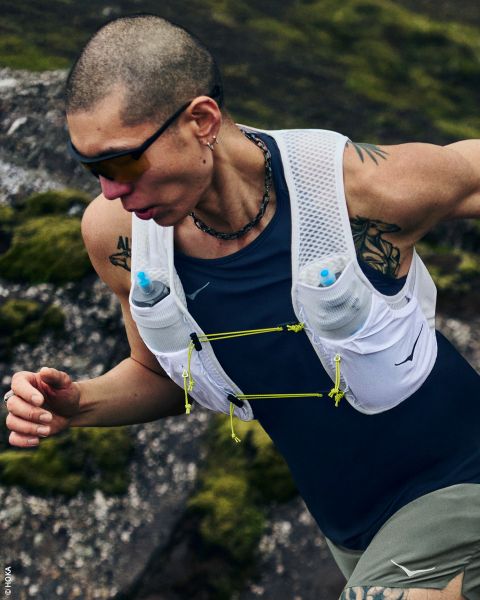 Une femme en tenue de sport et avec un bonnet porte un gilet de course avec des bouteilles d'eau et se tient de profil devant un simple fond blanc.