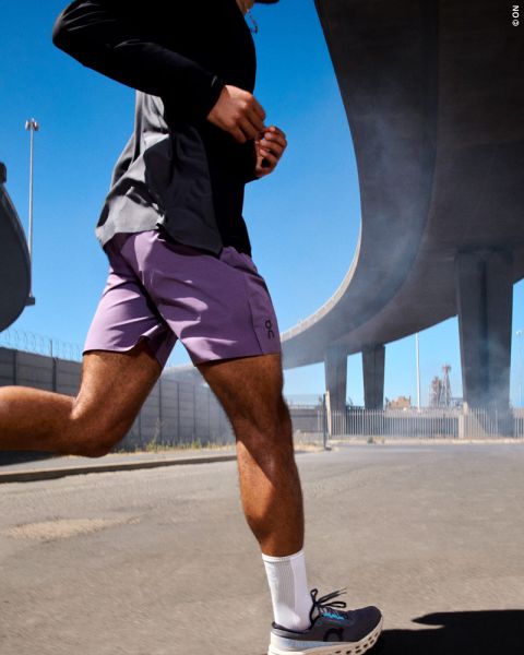 Une personne court à l'extérieur par une journée ensoleillée sous un grand pont, portant un short violet, une chemise noire, des chaussettes blanches et des chaussures de course.