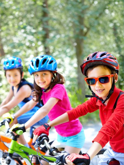 Trois enfants portant des casques font du vélo à l'extérieur par une journée ensoleillée et sourient à la caméra. L'arrière-plan est rempli d'arbres verts.