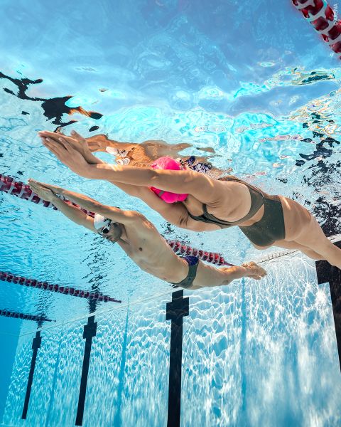 Deux nageurs, dont l'un porte un bonnet rose et l'autre un bonnet blanc, glissent côte à côte sous l'eau dans une piscine, nageant dans des couloirs séparés.