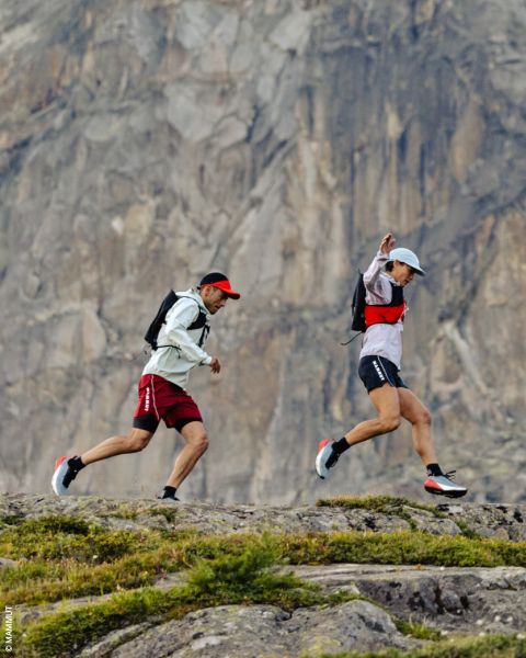 Deux personnes marchent sur un sentier rocheux devant une falaise de montagne abrupte, elles portent des chapeaux, des sacs à dos et des vêtements de sport.