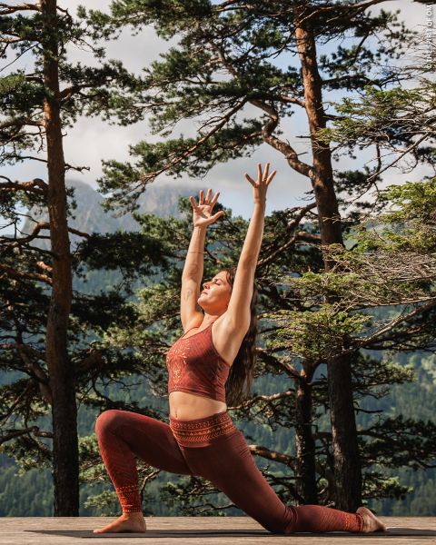 Une femme en tenue de sport rouge pratique le yoga en plein air sur une terrasse en bois, entourée de grands arbres et de montagnes, et étire ses bras au-dessus de sa tête dans une pose de fente.