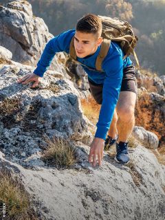 Un jeune homme en sweat-shirt bleu et avec un sac à dos escalade une pente rocheuse de montagne, entouré de feuillage d'automne.