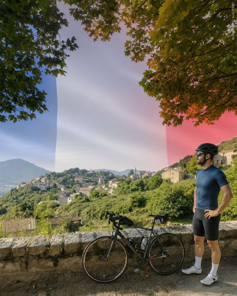 Un vélo est appuyé contre un mur de pierre, surplombant un village au milieu d'un paysage vallonné luxuriant, tandis que le drapeau français se découpe discrètement sur le ciel.