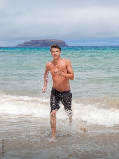 Ein hemdsärmeliger Mann mit Tattoos und schwarzen Badeshorts steht bei Sonnenuntergang im flachen Meerwasser und schaut in die Kamera. Hinter ihm rollen die Wellen heran.
