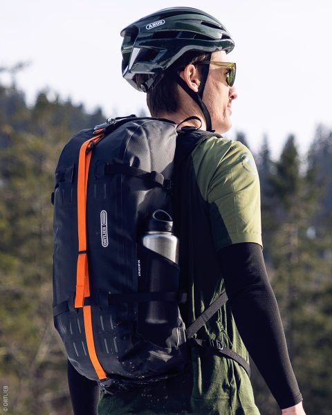 Une personne portant un casque vert et des lunettes de soleil, transportant un sac à dos noir avec une bande orange et une bouteille d'eau, se trouve à l'extérieur dans une zone boisée.
