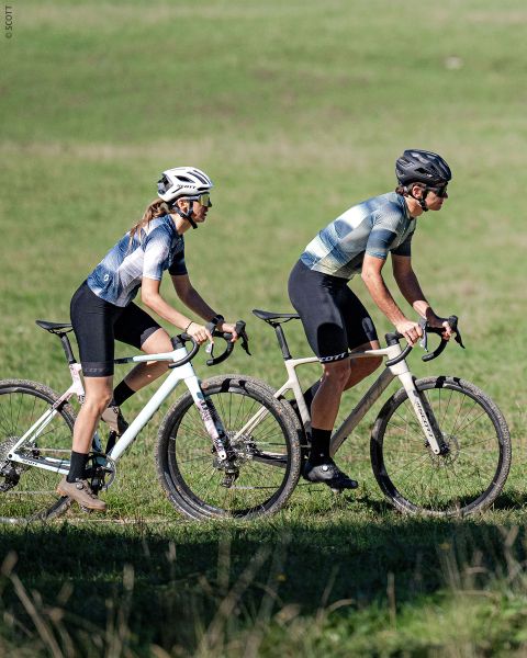 Deux cyclistes portant des casques et des vêtements de cyclisme roulent côte à côte sur des vélos blancs sur un chemin herbeux, avec une prairie verte en arrière-plan.