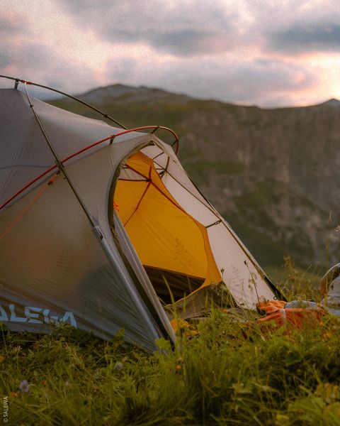 Une tente grise et jaune se dresse sur une prairie avec des montagnes en arrière-plan sous un ciel nuageux au coucher du soleil.