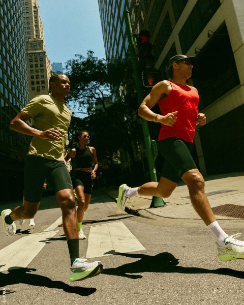 Trois personnes en tenue de sport et baskets courent dans une rue de la ville, avec de hauts bâtiments et la lumière du soleil en arrière-plan.