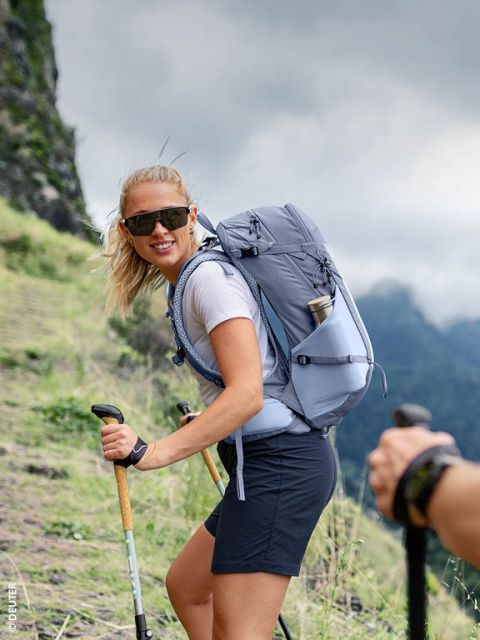Une femme portant des lunettes de soleil gravit un sentier de montagne herbeux avec des bâtons de marche et un sac à dos gris, tout en souriant à la caméra.