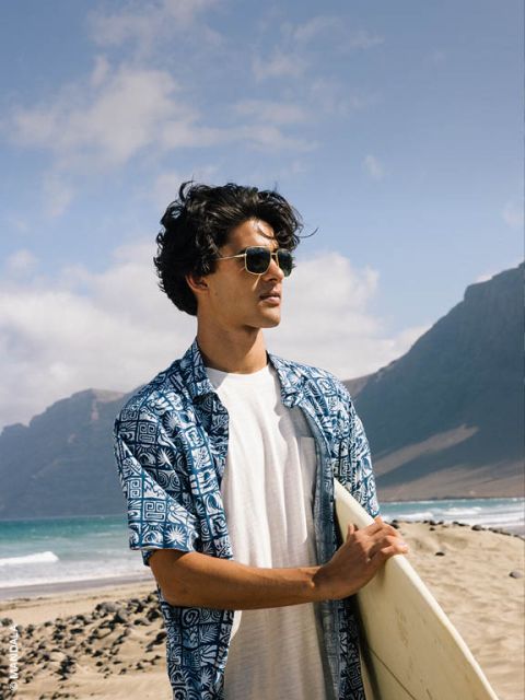 Un homme avec des lunettes de soleil et une chemise à motifs bleus tient une planche de surf et se tient sur une plage de sable avec des montagnes et l'océan en arrière-plan.