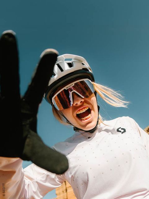 Un cycliste souriant portant un casque, des lunettes de soleil et des gants s'étire face à la caméra devant un ciel bleu éclatant.