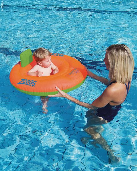 Une femme tient un bébé dans ses bras, qui est assis dans une bouée de natation orange et verte dans une piscine.
