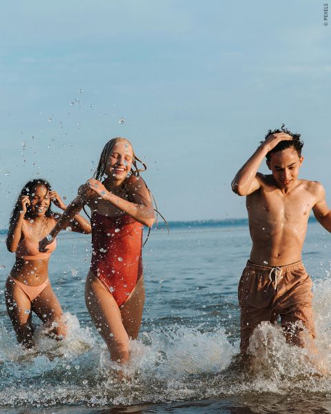 Trois jeunes adultes en maillot de bain rient et barbotent, tandis qu'ils marchent dans l'eau de mer peu profonde par une journée ensoleillée.