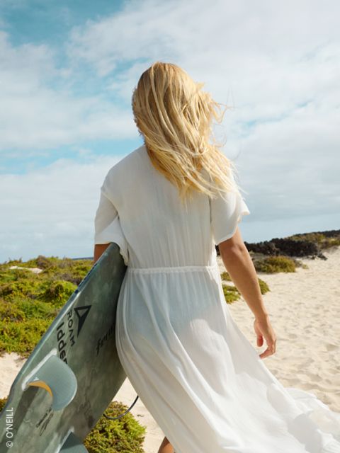Une femme en robe blanche marche avec une planche de surf à la main sur une plage de sable, ses cheveux blonds flottent au vent.