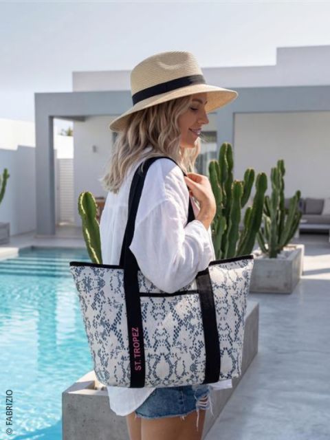 Une femme avec un chapeau de paille et une chemise blanche se tient au bord d'une piscine et tient un grand sac fourre-tout à motifs avec l'inscription "St. Tropez", avec des cactus en arrière-plan.