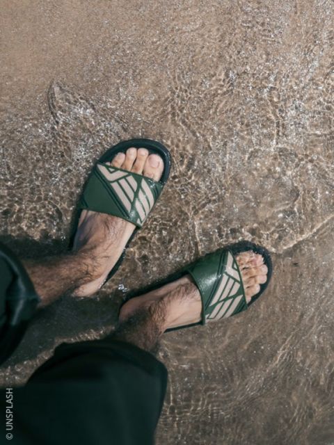 Une personne avec des sandales vertes se tient dans l'eau peu profonde sur une plage de sable, les jambes de son pantalon légèrement retroussées.