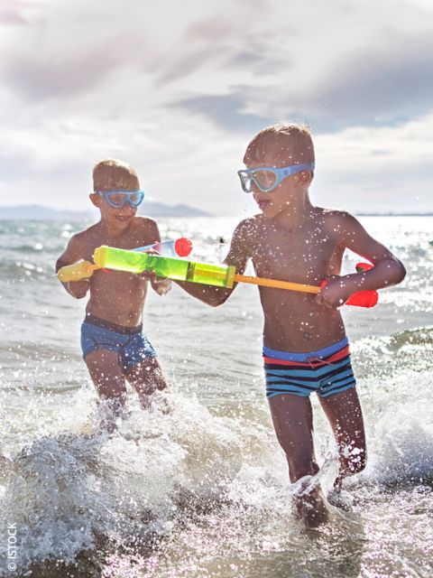 Deux garçons avec des lunettes de natation jouent avec un pistolet à eau dans la mer et barbotent dans les vagues sous un ciel clair et ensoleillé.