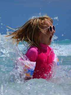 Une fille avec des lunettes de soleil roses et un maillot de bain barbotte dans la mer et sourit avec excitation, tandis que l'eau éclabousse autour d'elle par une journée ensoleillée.