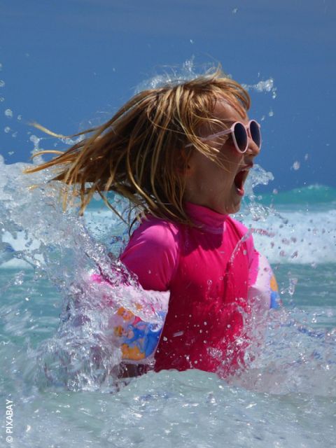 Une fille avec des lunettes de soleil roses et un maillot de bain barbotte dans la mer et sourit avec excitation, tandis que l'eau éclabousse autour d'elle par une journée ensoleillée.