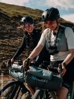 Deux femmes avec des casques et des lunettes de soleil roulent en souriant à vélo en plein air, avec des sacoches attachées. À l'arrière-plan, on peut voir une colline herbeuse.