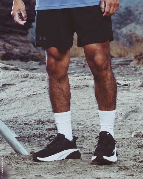 Une personne en shorts noirs, chaussettes blanches et baskets noires se tient sur un sentier de randonnée rocheux en plein air.