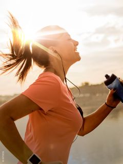 Une femme en t-shirt rose, faisant du jogging en plein air au lever du soleil, sourit, porte des écouteurs et tient une bouteille d'eau, tandis que le soleil brille derrière elle.