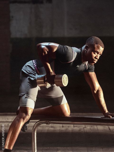 Un homme en tenue de sport soulève un haltère tout en s'appuyant sur un banc ; il s'entraîne concentré dans une salle de sport faiblement éclairée.