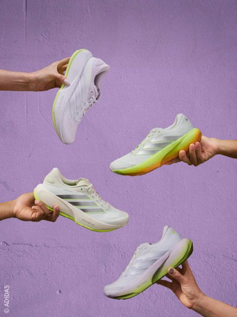 Quatre mains qui tiennent des baskets blanches avec des semelles vertes et jaunes devant un mur couleur lavande.