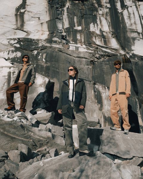 Trois personnes en tenue de plein air se tiennent sur un terrain rocheux, derrière elles un grand mur de pierre, et regardent au loin sous un soleil éclatant.