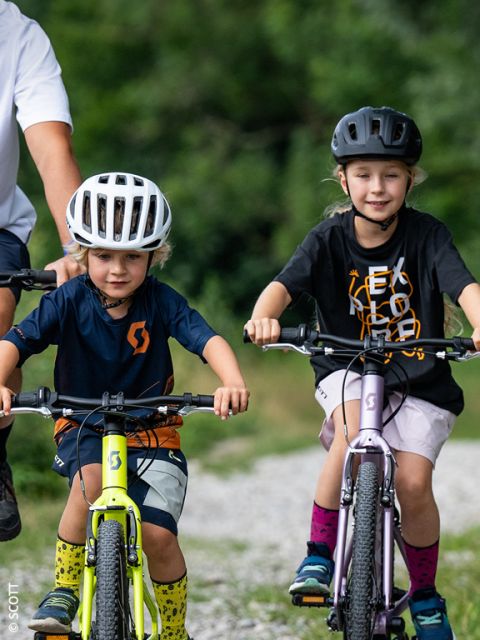 Deux petits enfants avec des casques font du vélo à l'extérieur sur un chemin de gravier, tandis qu'un adulte se tient à proximité. De la verdure est visible en arrière-plan flou.