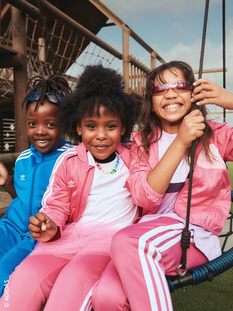 Trois enfants souriants en tenues Adidas sont assis dehors sur une balançoire - deux d'entre eux en survêtements roses et un en un bleu - et profitent de la journée ensoleillée.