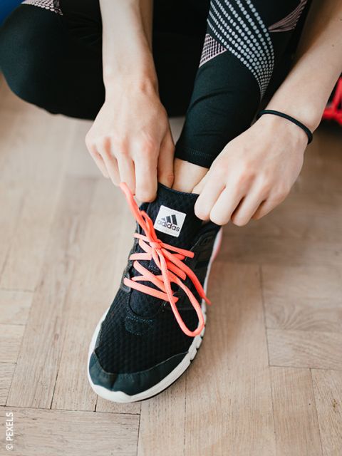 Une personne qui noue des lacets rose vif sur une basket Adidas noire, et porte des leggings noirs avec des motifs blancs et roses.