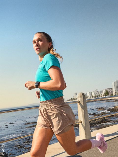 Une femme en tenue de sport fait son jogging le long de la mer par une journée ensoleillée, avec les bâtiments de la ville visibles en arrière-plan.