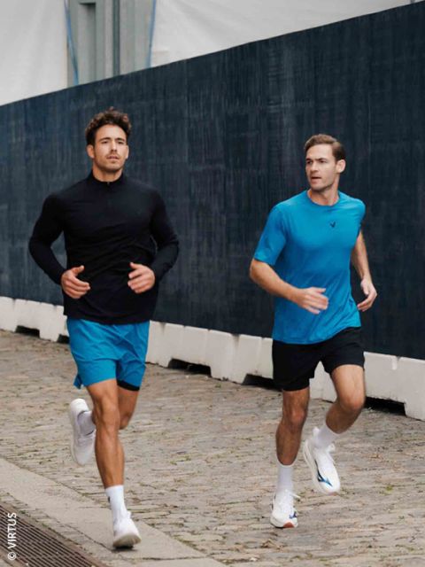 Deux hommes en tenue de sport font du jogging sur un chemin pavé à côté d'une clôture sombre ; tous deux portent des shorts bleus et des baskets blanches.