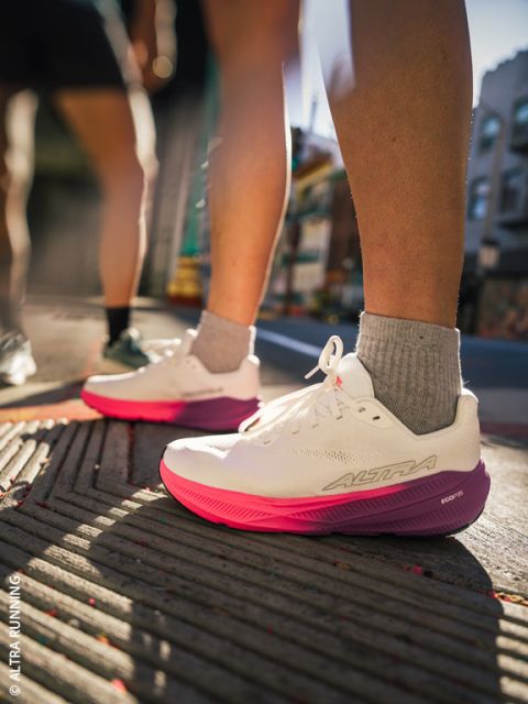 Une personne en chaussures de course blanches et roses se tient sur un trottoir en ville ; la lumière du soleil fait ressortir les chaussures et les jambes, tandis que l'arrière-plan urbain semble flou.