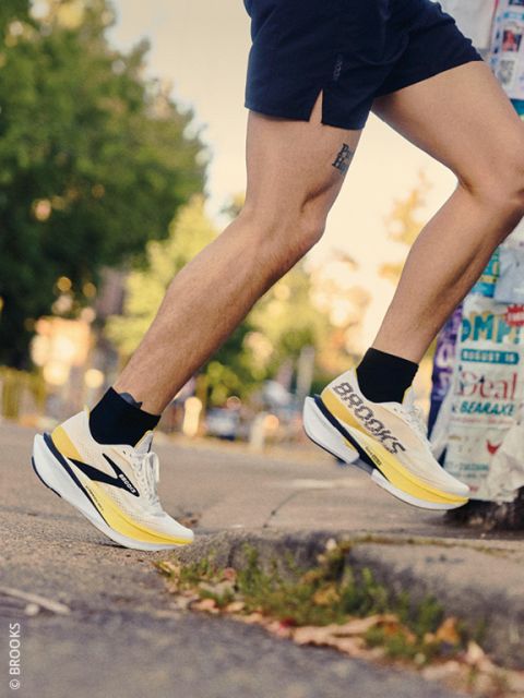 Un coureur en short et des chaussures de course Brooks jaunes et blanches monte sur un trottoir. À l'arrière-plan, on peut voir des arbres et un poteau recouvert d'affiches.