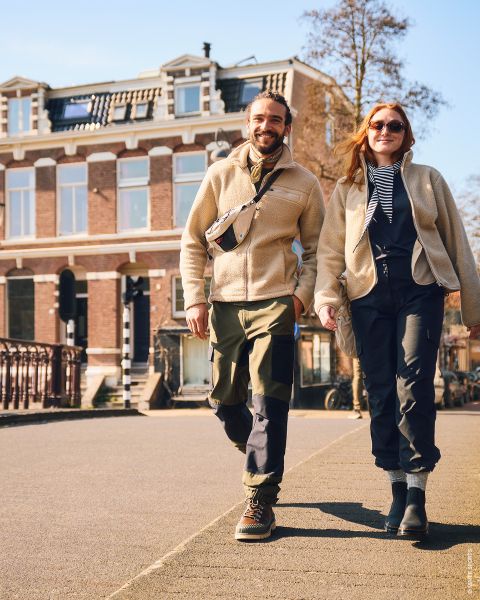 Un homme et une femme souriants se promènent en plein air par une journée ensoleillée, vêtus de vestes et de bottes décontractées, se tenant la main, avec un grand bâtiment en briques en arrière-plan.